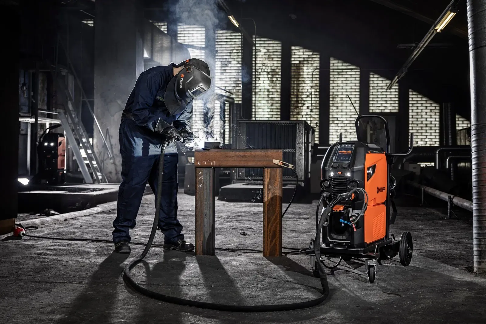 A welder working at a steel table with a Master M behind
