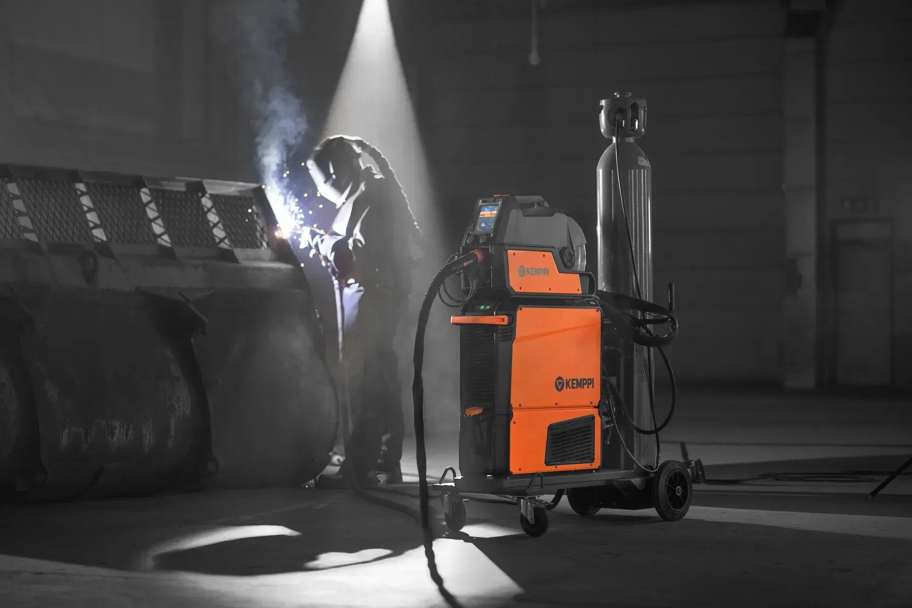 A welder working in a dark hall beside an X5 FastMig on a trolley